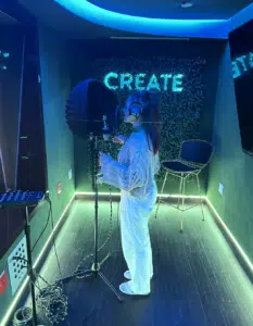 A person wearing headphones stands at a microphone in a recording studio with "CREATE" illuminated on the wall behind them.